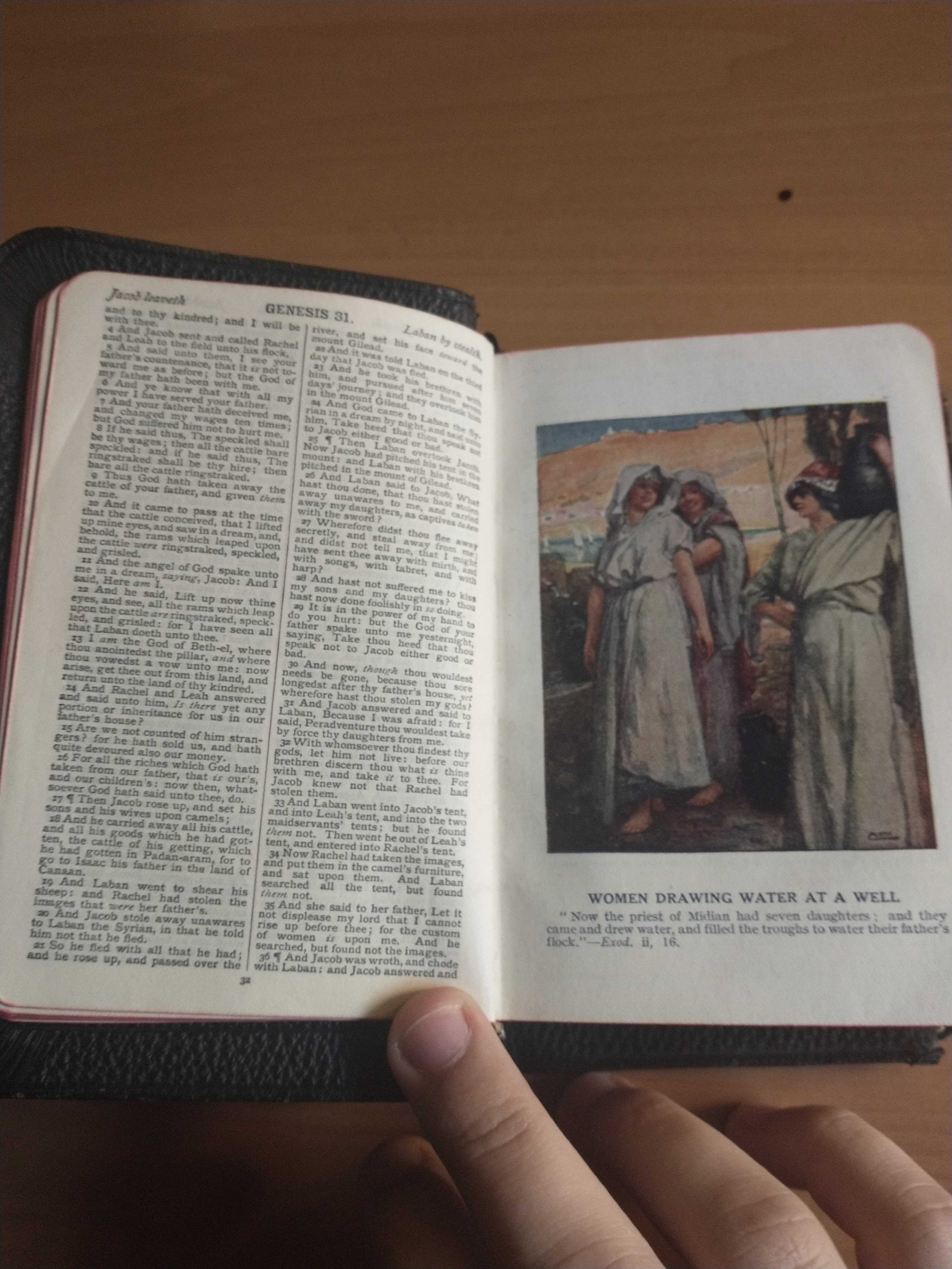 The Bible is open to pages 31 and 32. You can see that the pages have a red outline on their edges. On the left page is Genesis 31, in two columns. The text is very small. On the right page is an illustration titled "Women Drawing Water At A Well". It is in the same style as the previous illustration. It depicts three women it loose white robes, smiling and carrying water. There are plants around them. In the background appears to be a small pond, and a desert hill.