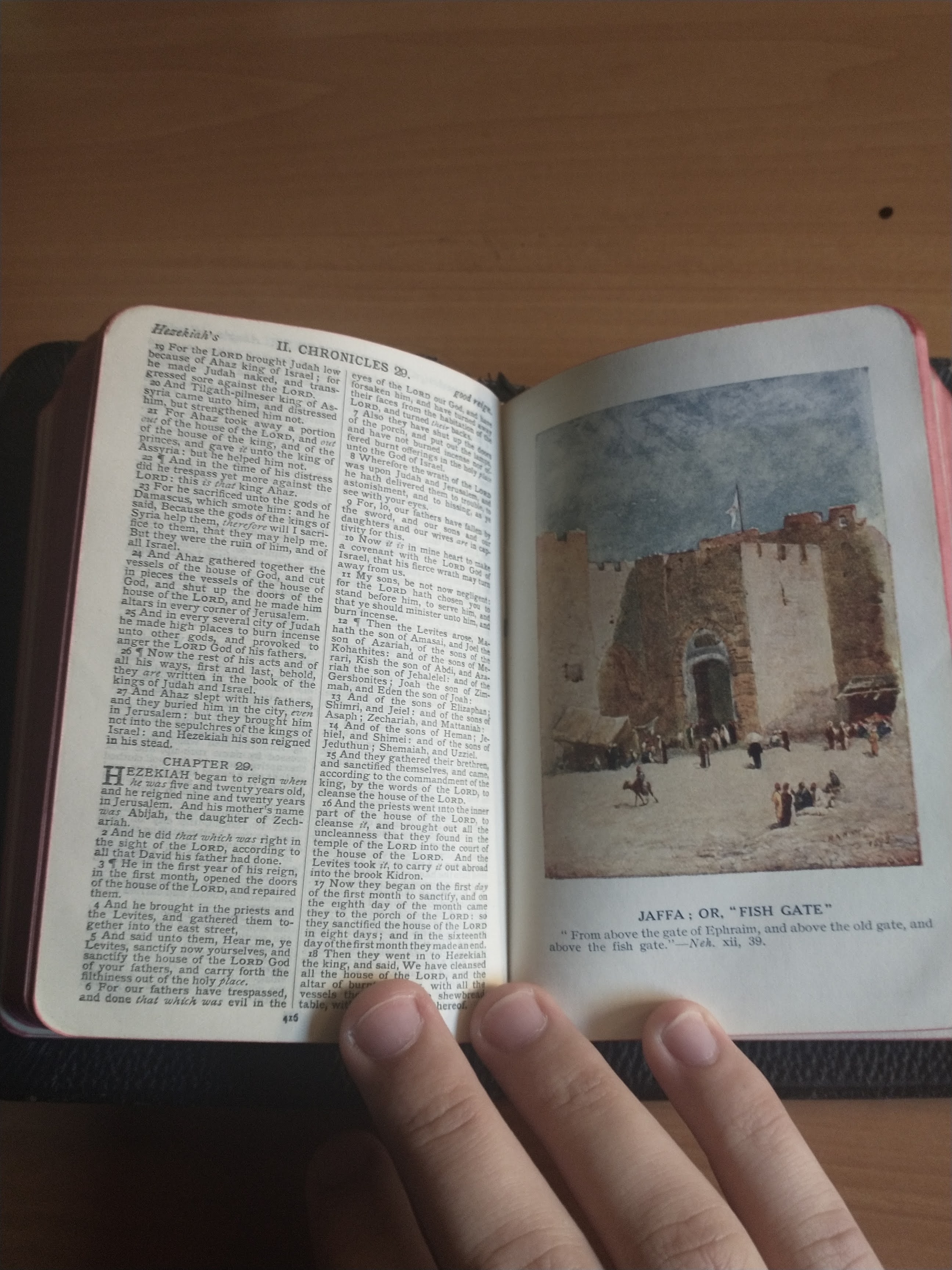 The Bible is open to pages further into the book. On the left page is II. Chronicles 29, in the same style as the previous photo. On the right page is an illustration titled "Jaffa; Or, "Fish Gate"". It is in the same style as before. In the foreground is a big sandstone castle with a white flag above it. There is an open gate, and people are walking in and out, and gathering around. There are two dilapidated market stands against the walls of the castle. A man travels by on a camel. The illustration is detailed enough to make out that these are people, and to see generally what they are doing, but not enough to make out any more personal details. The sky is blue.